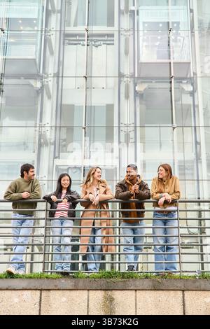 Five young students chatting happily on a balcony of a modern building, enjoying their break and sharing laughter against a vibrant cityscape Stock Photo