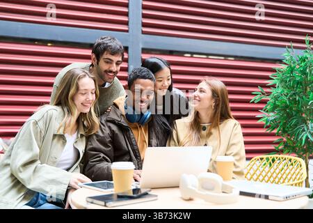 Five cheerful young multiethnic students are using a laptop outside, enjoying their time together while working on a project Stock Photo