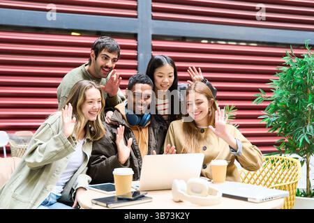 Group of cheerful diverse students waving at laptop making video call sitting at table in outdoor cafe Stock Photo