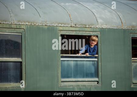 Blond boy leans out train window study rural tracks and fields Stock Photo