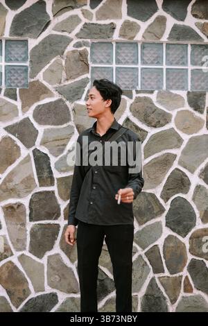Young man in black attire standing by a stone wall while holding a cigarette Stock Photo