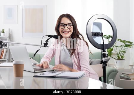 Smiling young female content creator and influencer at home, using smartphone and ring light, working with laptop and microphone. Live streaming or re Stock Photo
