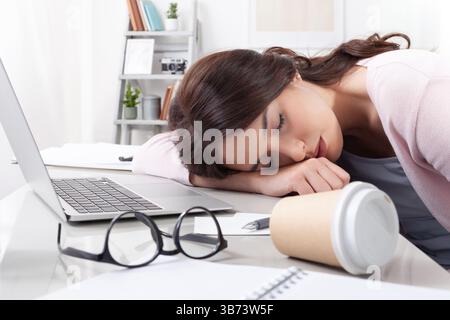 Overworked and exhausted young female student or freelancer, sleeping on her desk after long hours of work, study, or hours spent online, surrounded b Stock Photo