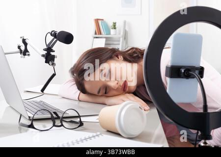 Exhausted female content creator and influencer asleep at desk with laptop, smartphone, and ring light. Tired and overworked from recording video tuto Stock Photo