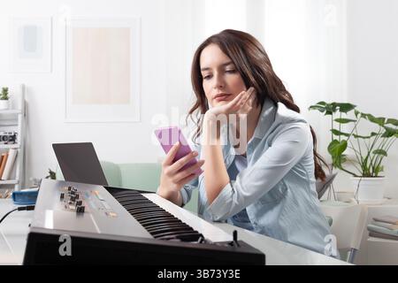 Young woman at home, playing music keyboard and use smartphone, learning music, composing, searching online, or shopping. Concept of creative work, dr Stock Photo