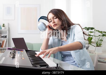 Young woman at home, playing a music keyboard while using headphones, laptop, and a smartphone. Learning and composing music. A concept of creative wo Stock Photo