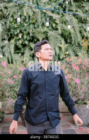 Man looks pensively upwards in a lush, green garden with decorative string lights Stock Photo