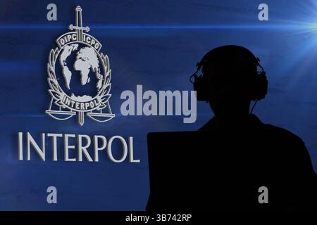 Silhouette of a man wearing headphones in front of a laptop against the background of the Interpol logo symbolising cybersecurity and surveillance Stock Photo