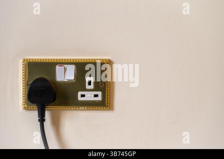 UK style double electrical socket with plug inserted. Gold socket with white details, black plug and magnolia wall. One switch in on position. Stock Photo