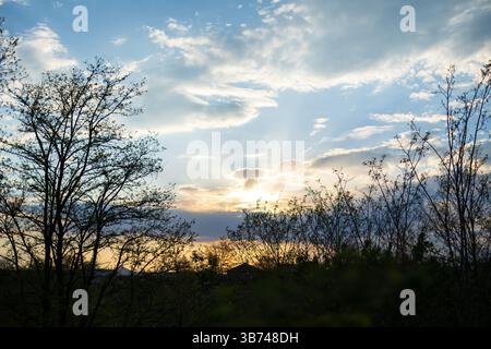 The sun is setting behind hills with trees. The sky is clear blue, dotted with clouds. Stock Photo