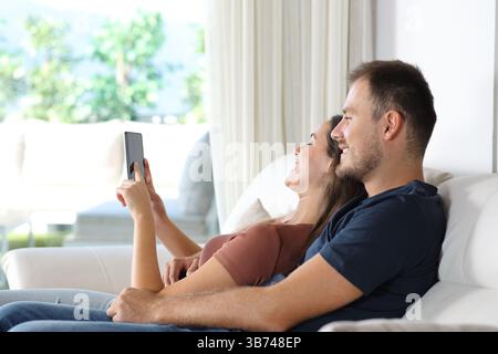 Happy couple using smart phone sitting on a couch in a luxury house Stock Photo