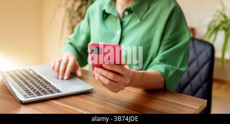 Woman using smartphone with pink case at the desk with laptop. Person working from home. Stock Photo