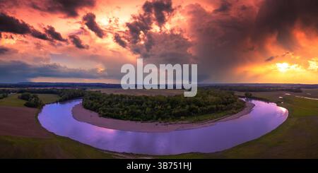 A vibrant sunset with a faint funnel cloud and distant storm over a dramatic bend in the white river. Blue purple water, orange red skies. Green trees Stock Photo