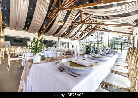 Wedding setting and decorated tables in a beautiful modern interior of a performance hall with a modern light ceiling of wooden construction. Stock Photo
