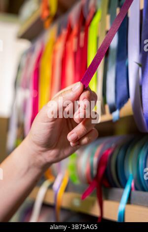This vertically oriented close-up captures a hand gently holding a purple ribbon from a display rack filled with multicolored satin ribbons Stock Photo