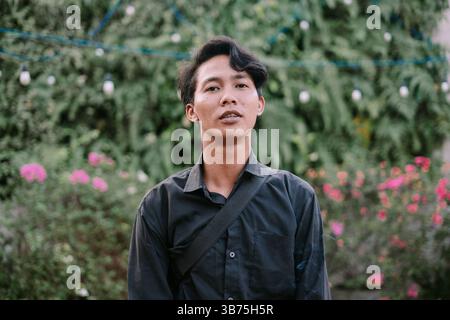 Authentic portrait of a young Asian man. He stands confidently amidst a green garden backdrop with pink flora Stock Photo