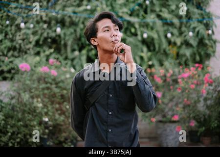 Young man holding a cigarette, taking a contemplative moment amidst lush greenery outdoors. Casual style with a thoughtful expression Stock Photo
