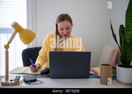 Woman in yellow sweater works on laptop and writes notes at a home office desk with lamp and plant  Stock Photo