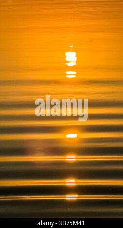 Golden sunset reflections over calm water with radiant ripples creating a serene and peaceful atmosphere amidst a warm orange glow. Stock Photo