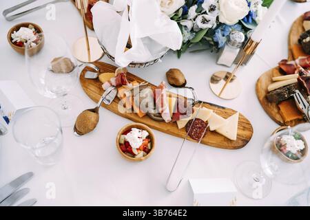 A wooden cutting board with pieces of delicacies on a white tablecloth in the center of the festive table. Stock Photo