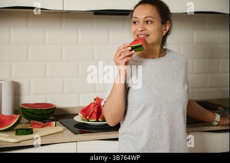 A cheerful moment captured as a woman enjoys a slice of refreshing watermelon in a cozy kitchen. The setting showcases a healthy, relaxed lifestyle wi Stock Photo