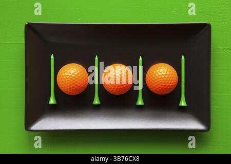 Black ceramic dishes with golf balls and wooden tees on over green background, rectangle dish Stock Photo