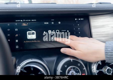 A woman's hand turns up the sound volume on the dashboard in a modern car. The Car Dashboard features a modern Touchscreen integrated with a Radio Sta Stock Photo