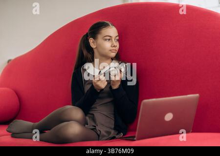 Thoughtful girl taking break during online lesson sitting on red couch holding glasses near laptop showing moment of fatigue and reflection in digital Stock Photo