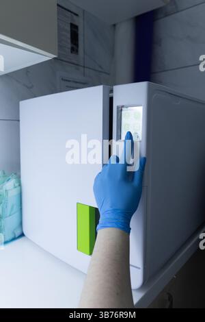 This image shows a hand wearing a blue glove pressing a button on a sleek, modern refrigerator's digital interface in a clean, clinical setting, sugge Stock Photo