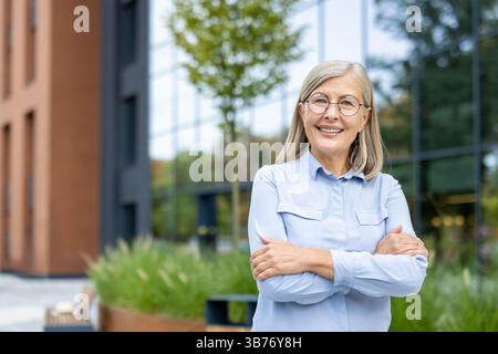 A smiling senior woman with crossed arms poses outdoors near a modern building with glass windows. Stock Photo