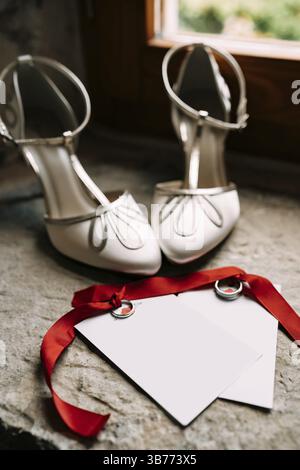 Wedding rings tied with red ribbons lie on blank postcards with ornaments next to white high-heeled shoes against the background of a stone window sil Stock Photo