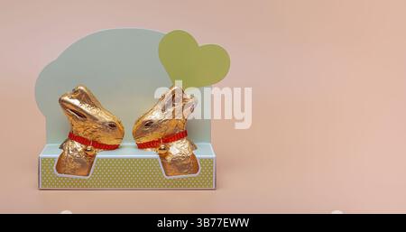 Two golden chocolate Easter bunnies wrapped in foil with red ribbons, displayed on a pastel backdrop with a green heart Stock Photo