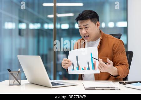 An asian businessman gestures towards a chart during a video conference presentation, using his laptop and a pen to illustrate points Stock Photo