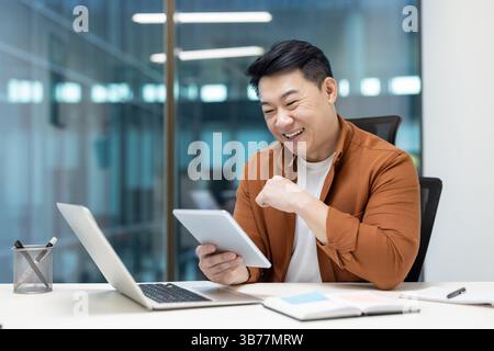 A cheerful programmer enjoys testing an application on a tablet, seated at a modern office desk with a laptop Stock Photo