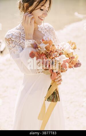Beautiful bride in a white dress with sleeves and lace, with a yellow autumn bouquet of dried flowers and peony roses, on the Lago di Braies in Italy. Stock Photo
