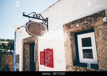 Close-up of a rustic blank hanging shop sign mockup on a traditional whitewashed building with stone framing, red shuttered menu window, and vintage l Stock Photo