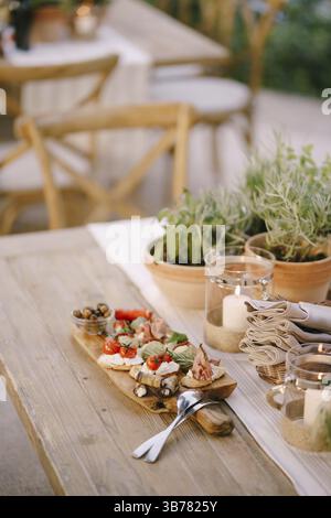 Wooden cutting board with cold cuts and cutlery on the table by the flowers in the pots. Stock Photo