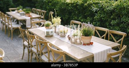Wedding dinner table reception at sunset outside. Ancient rectangular wooden tables with rag runner, wooden vintage chairs, lavender pots, cherry toma Stock Photo