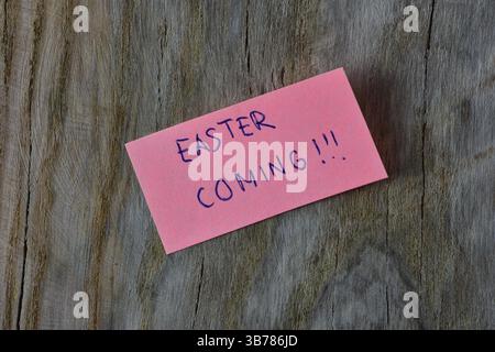 Words easter coming on pink sticky note on wooden board textured background. A pen wrote words easter coming on note paper. Stock Photo