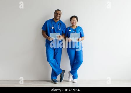 Two african american healthcare professionals in blue scrubs, leaning against white wall Stock Photo