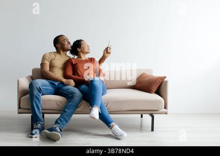 Split system. Cheerful young black couple opening air conditioner with remote Stock Photo