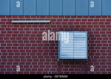 An air conditioning unit is mounted on a red brick wall beneath a blue roof. The unit features metallic, slatted coverings and is positioned away from Stock Photo