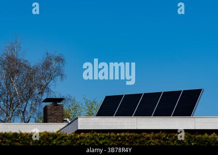 A row of solar panels is installed on a flat roof, with a chimney visible nearby. The sky is bright and blue, suggesting a sunny day perfect for harne Stock Photo