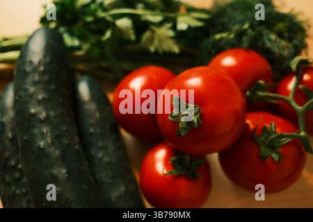 Fresh red tomatoes on the vine, dark green cucumbers, and sprigs of parsley and dill arranged on wooden table. A rustic, vibrant still life highlighti Stock Photo