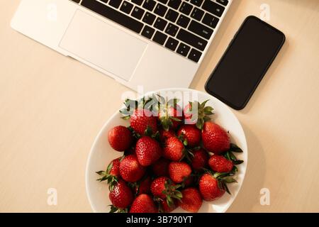 A white bowl of glossy red strawberries sits beside a sleek smartphone and silver laptop on a light wood desk, blending fresh produce with modern tech Stock Photo