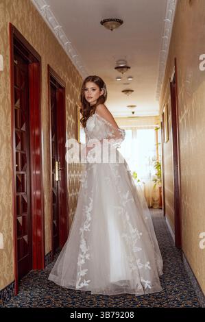 Young bride in a long classic dress in the hotel corridor. On the silk dress lace Stock Photo