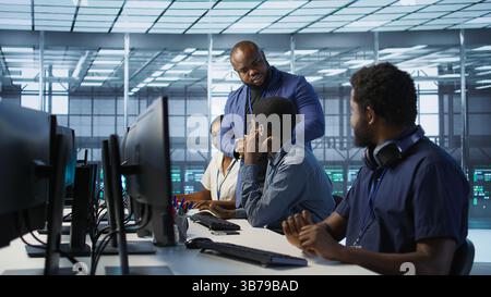 Data center team leader supervising engineers implementing AI predictive maintenance systems. Server farm boss overseeing workers using artificial intelligence to identify hardware failures, camera A Stock Photo
