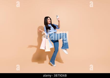 Young woman happily poses with shopping bags and takes a selfie against a beige background in casual stylish attire Stock Photo