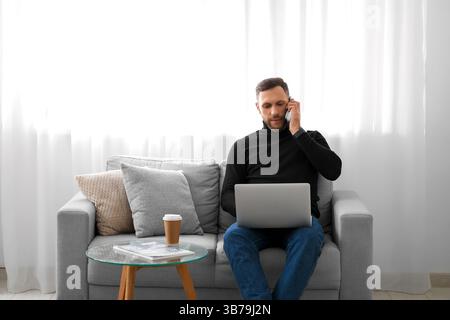 Young freelancer with laptop talking on mobile phone at home Stock Photo
