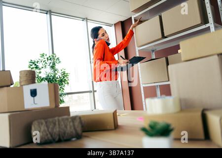 Professional young businesswoman managing orders in a busy workspace while checking documents and organizing boxes indoors. Stock Photo